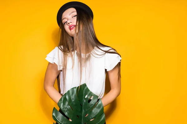 Modelo de menina de cabelos compridos luxuoso em um chapéu na moda preto detém uma folha verde e posa em um fundo laranja — Fotografia de Stock