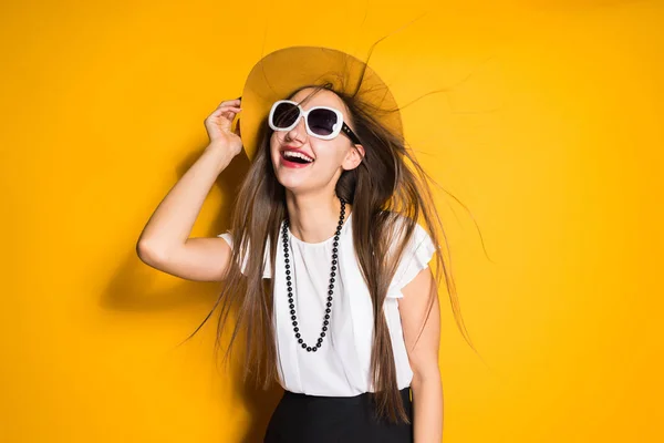 Улыбающаяся длинноволосая модель в шляпе и солнцезащитных очках, позирующая на желтом фоне — стоковое фото