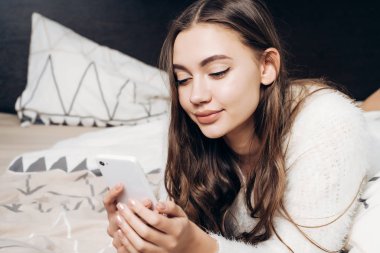 Sevimli güzel kız yatakta, dinlenme, gülümseyen ve onun smartphone bakarak yalan pijama