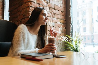 gülümseyen güzel uzun saçlı kız öğrenci bir beyaz kazaklı bir kafede oturuyor, lezzetli kahve içer ve pencereden görünüyor