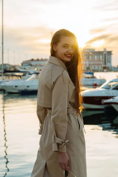 Feliz chica de pelo largo de lujo en un abrigo beige posando en el sol de la noche en el puerto — Foto de Stock