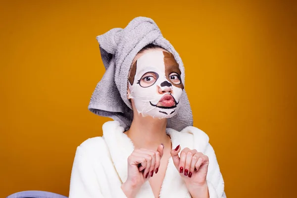 滑稽的年轻女孩用毛巾在她的头上照顾自己, 在她的脸上有一个带有狗脸的保湿面具 — 图库照片