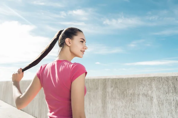 Selbstbewusstes schlankes langhaariges Mädchen in rosa T-Shirt, das sich vor dem Training an der frischen Luft ausruht und Kraft tankt — Stockfoto