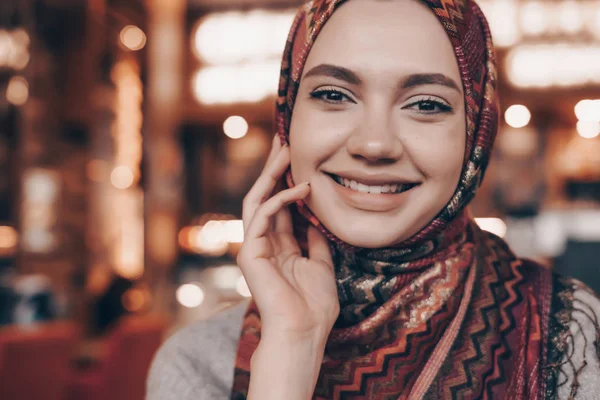 Hermosa chica musulmana sonriente con un pañuelo en la cabeza sentado en un café y esperando su comida — Foto de Stock