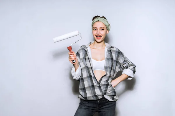Смешная молодая девушка в клетчатой рубашке окрашивает стены в белый цвет с роликом, ремонт в квартире — стоковое фото