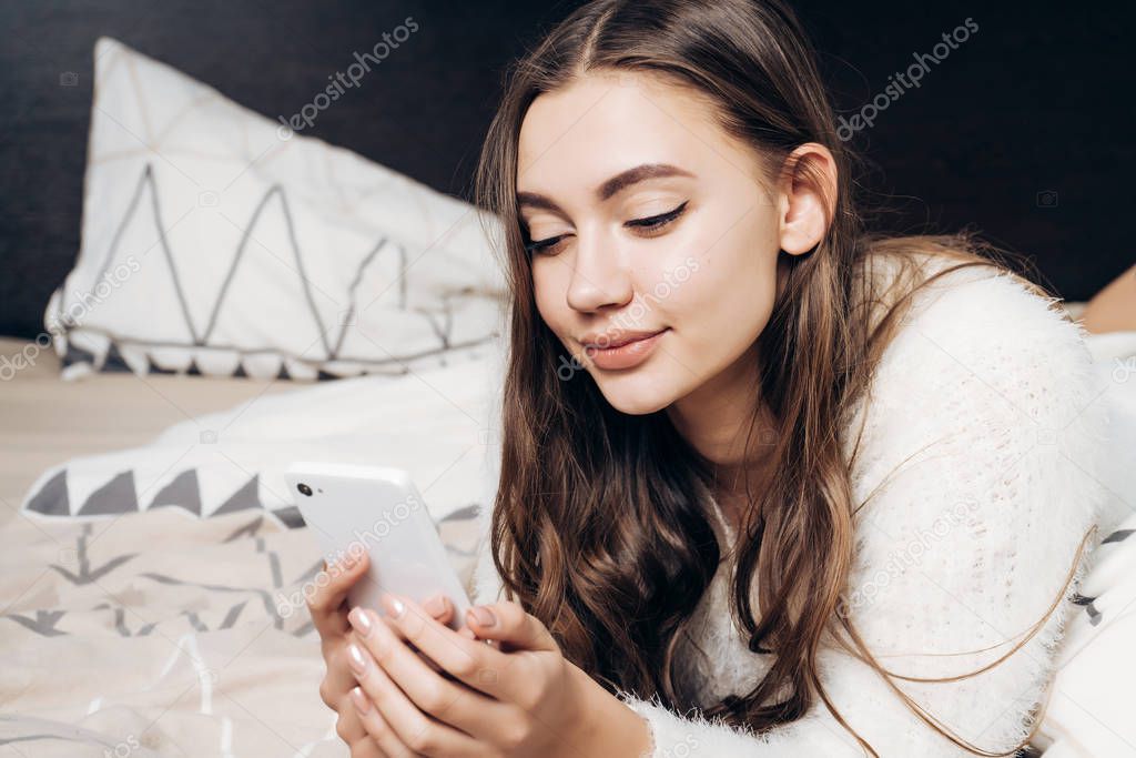 cute beautiful girl in pajamas lying on bed, resting, smiling and looking into her smartphone