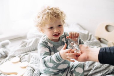 Anne, İçecek su ve kahkaha ile küçük kıvırcık oğlan mavi pijama yatağa oturur