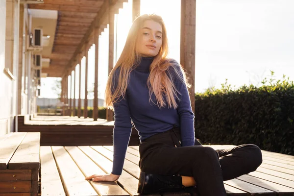 Bonito jovem loira menina em camisola azul senta-se em um banco de madeira no parque e posa — Fotografia de Stock