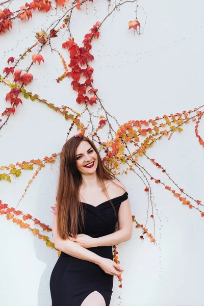 Luxurious long-haired girl in a black dress posing against the wall in her garden, smiling and resting — Stock Photo, Image