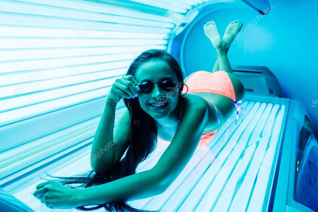 attractive slender girl in a bathing suit sunbathing in a horizontal tanning bed, wearing safety glasses, smiling