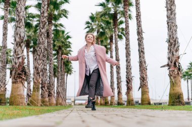 uzun zaman pembe bir ceket neşeli genç kadın uzun boylu yeşil palms satırlarla bir parkta yürüyor