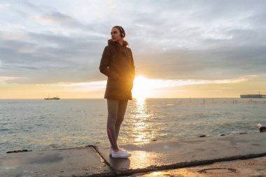 kulaklıklar müzik dinlemek gün batımında, deniz sahil boyunca sıcak bir ceketi giyen genç bir kız yürüyor