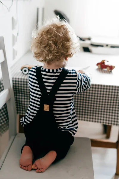 Malý kudrnatý chlapeček v roztomilé overaly sedí na židli v kuchyni a kreslení — Stock fotografie