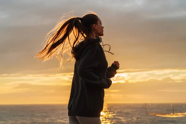 Corridore attivo dai capelli lunghi che corre in riva al mare al tramonto, preparandosi per una maratona — Foto Stock