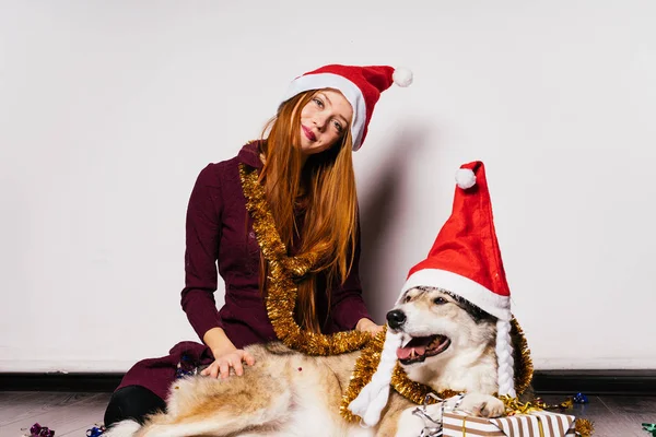 一个甜美的红头发的女孩, 像圣诞老人的帽子, 正在等待一个新的一年和圣诞节, 与她的大狗 — 图库照片