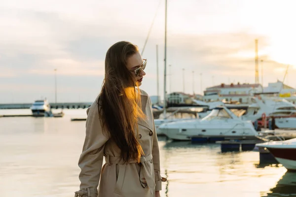 Luksusowa dziewczyna długowłosy w beżowy płaszcz czeka na jej jachtu morskiego, pozowanie na zachodzie słońca — Zdjęcie stockowe