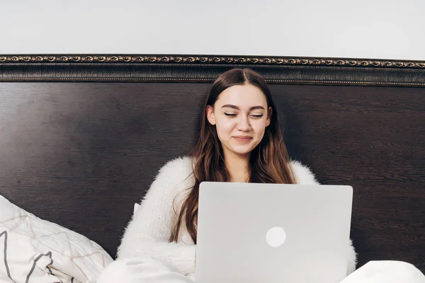 Красивая улыбающаяся девушка в пижаме лежит в постели вечером, смотрит смешное кино на ноутбуке — стоковое фото