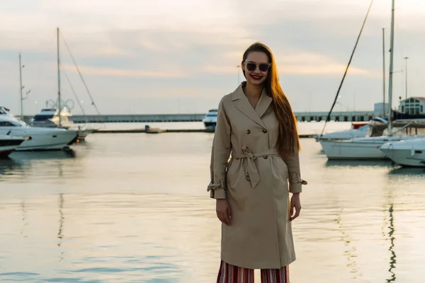 Bela menina de cabelos longos elegante em óculos de sol posando ao lado do mar à noite, sorrindo — Fotografia de Stock