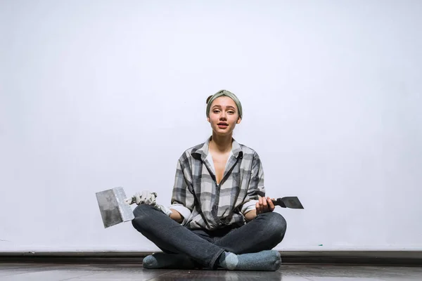 Een Moe meisje in een plaid shirt zit op de vloer in haar nieuwe appartement, spatels houdt in haar handen, reparaties — Stockfoto