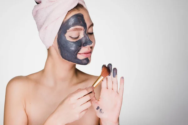 Okouzlující mladá dívka s ručníkem na hlavě aplikuje masku užitečné hlíny na polovinu tváře s kartáčem — Stock fotografie