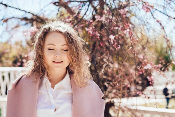 Charming curly young woman in a pink coat enjoying the warmth and sun in the park, closed her eyes — Stock Photo, Image