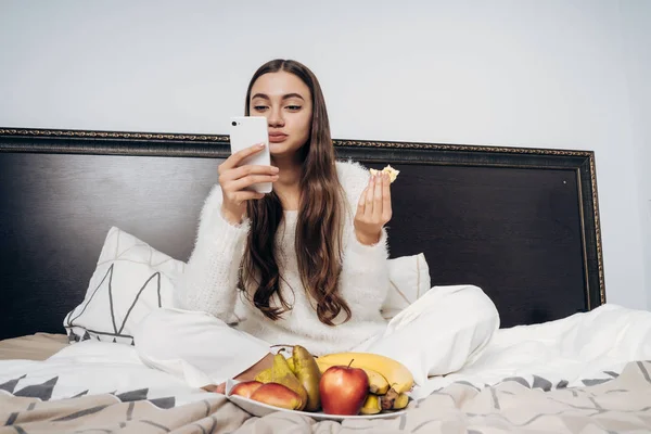 Мила довгошерста дівчина в білій піжамі сидить в ліжку рано вранці, їсть фрукти і дивиться в її смартфон — стокове фото