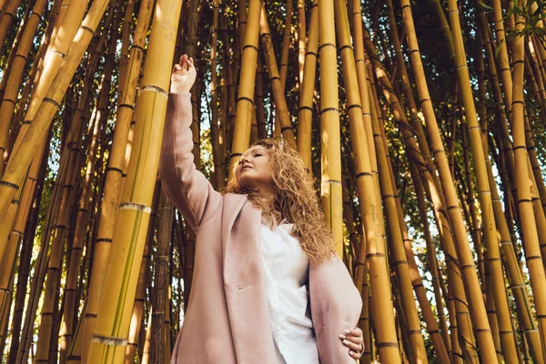 Stylish curly woman in a pink coat enjoying the nature in the park, next to the tall bamboo trees — Stock Photo, Image
