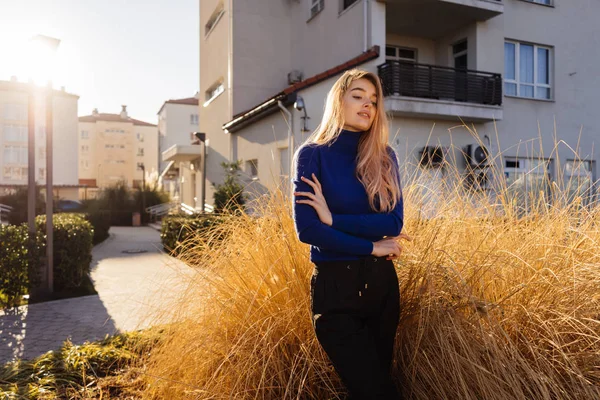 Роскошная блондинка в длинном голубом свитере позирует на солнце на улицах города, улыбаясь — стоковое фото