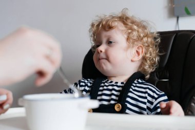 Mutfakta küçük bir Kıvırcık sarışın çocuk oturur, Anne beslemek lezzetli bir çorba, bir aile yemeği