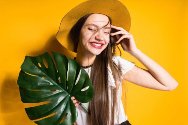 sarı arka plan üzerinde poz, yeşil yaprak tutan ve gülümseyen moda şapkalı güzel uzun saçlı kız modeli