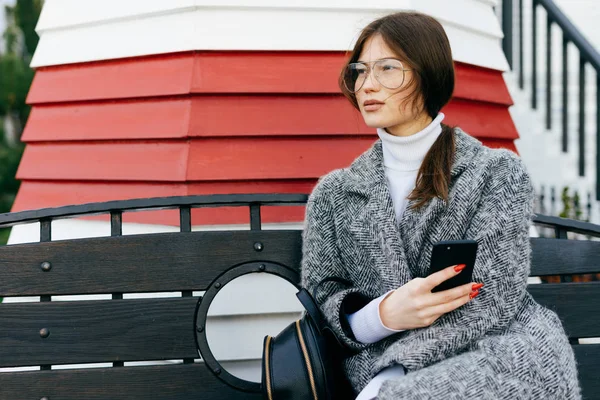 Élégante fille aux cheveux bruns avec des lunettes et un manteau gris reposant sur un banc à l'extérieur, tenant un smartphone — Photo