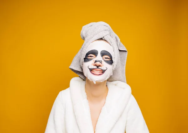 Een leuk meisje met een handdoek op haar hoofd en een witte badjas jurk ziet er na zichzelf, op haar gezicht een hydraterende masker met een gezicht van de panda — Stockfoto