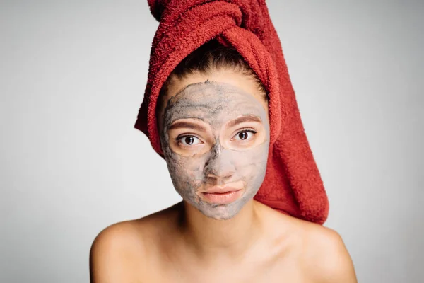 Schönes junges Mädchen mit rotem Handtuch auf dem Kopf trägt eine nützliche Tonmaske auf ihr Gesicht auf, blickt in die Kamera — Stockfoto