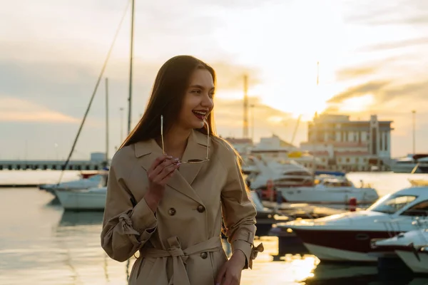 해질녘 항구에서 포즈 유행 베이지색 코트에 아름 다운 긴 머리 웃는 소녀 — 스톡 사진