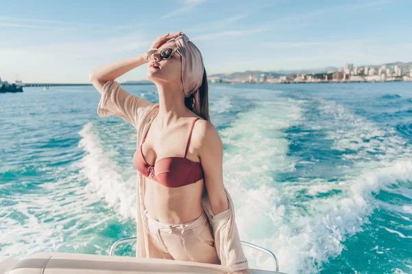 Hermosa mujer delgada con un pañuelo en la cabeza posando en un barco al sol, disfrutando de unas vacaciones y una vida rica —  Fotos de Stock