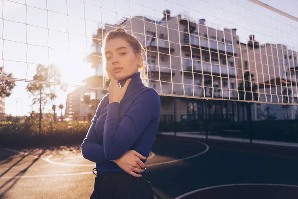 Впевнена стильна дівчина блондинка в синій куртці позує на спортивному полі в променях ранкового сонця — стокове фото