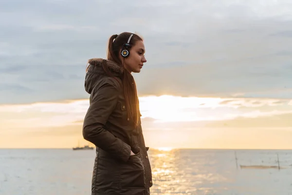 Carina ragazza ascoltando musica sulle cuffie, passeggiando al mare la sera — Foto Stock