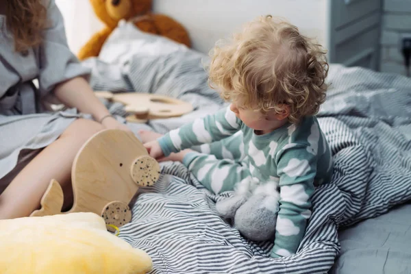 Malé kudrnaté blond chlapeček v modrém pyžamu sedí na posteli vedle jeho máma — Stock fotografie