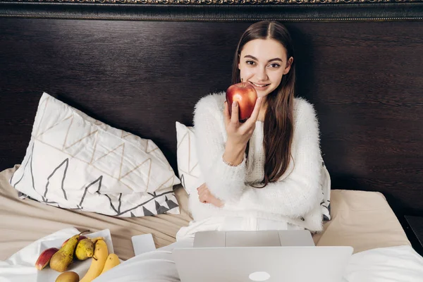 Красива усміхнена дівчина сидить в ліжку рано вранці, їсть яблуко і посміхається, з ноутбуком — стокове фото