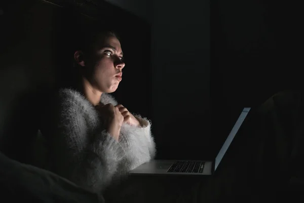 Menina assustada sentada na cama tarde da noite, assistindo filme de terror em seu laptop, no escuro — Fotografia de Stock