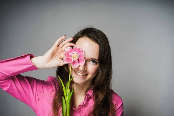 Menina bonita em uma camisa rosa segura uma flor pelo rosto, olha para a câmera e sorri — Fotografia de Stock