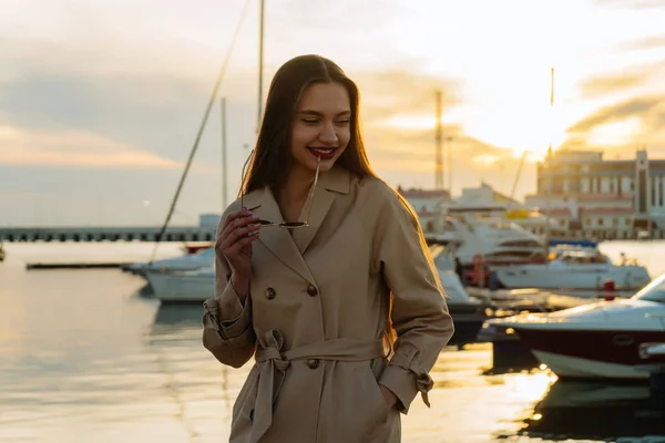 Attractive long-haired girl in fashionable beige coat posing in seaport at sunset, smiling, enjoying vacation — Stock Photo, Image