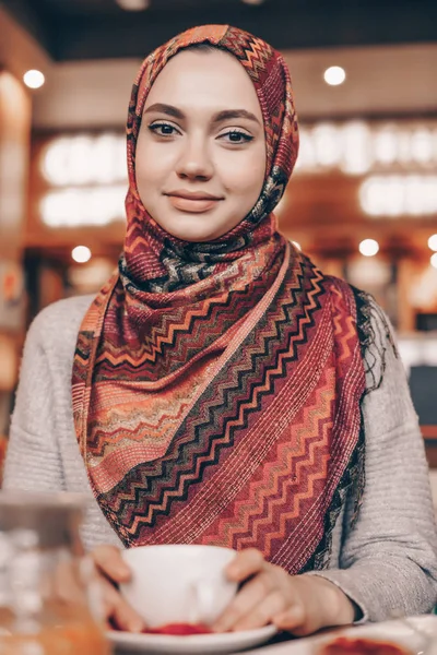 Charmantes arabisches Mädchen mit schönem Kopftuch beim Abendessen in einem gemütlichen Café, blickt in die Kamera und lächelt — Stockfoto