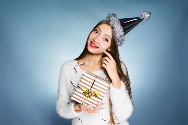 Feliz joven con una gorra festiva en la cabeza sostiene un regalo de Año Nuevo en sus manos y sonríe —  Fotos de Stock