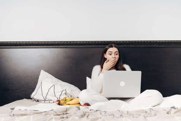 Sorprendida joven en pijama descansando en la cama comiendo fruta y viendo una película de miedo en su computadora portátil — Foto de Stock