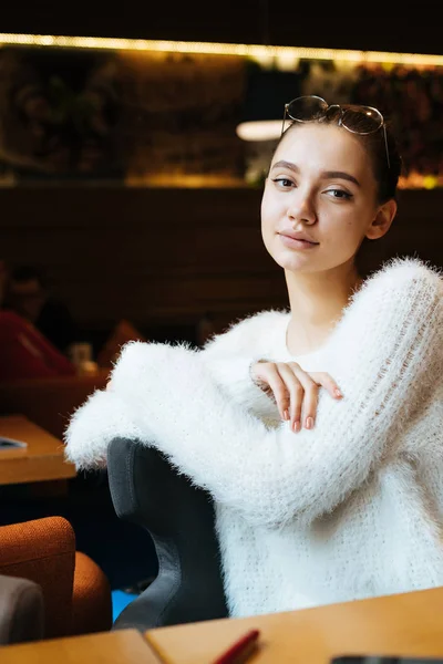 Задумчивая юная студентка в белой куртке отдыхает после учебы в кафе, смотрит в камеру — стоковое фото