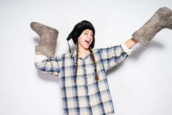 Divertida chica rusa con coletas en un sombrero de piel caliente posando y sonriendo, sosteniendo botas de fieltro en las manos — Foto de Stock