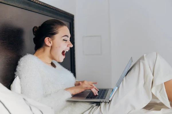 Alegre risueño chica en blanco pijama sentado en cama tarde en la noche, viendo divertido vídeo en su portátil —  Fotos de Stock