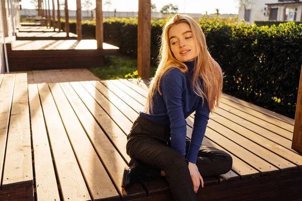 Atraente jovem loira modelo menina na moda camisola azul senta-se em um banco de madeira no parque, sorrindo — Fotografia de Stock