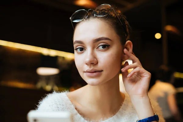 Красива молода дівчина студентка в білому светрі сидить в кафе перед заняттям, дивиться на камеру — стокове фото